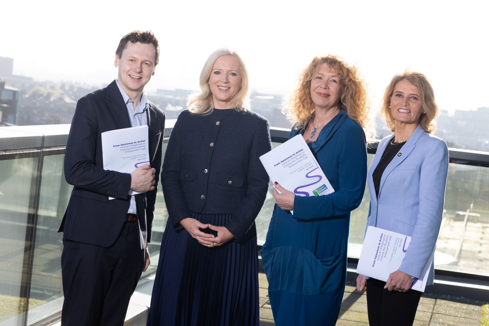 Four smiling people in business dress standing on a sunny balcony, three women and one man, holding copies of a printed report