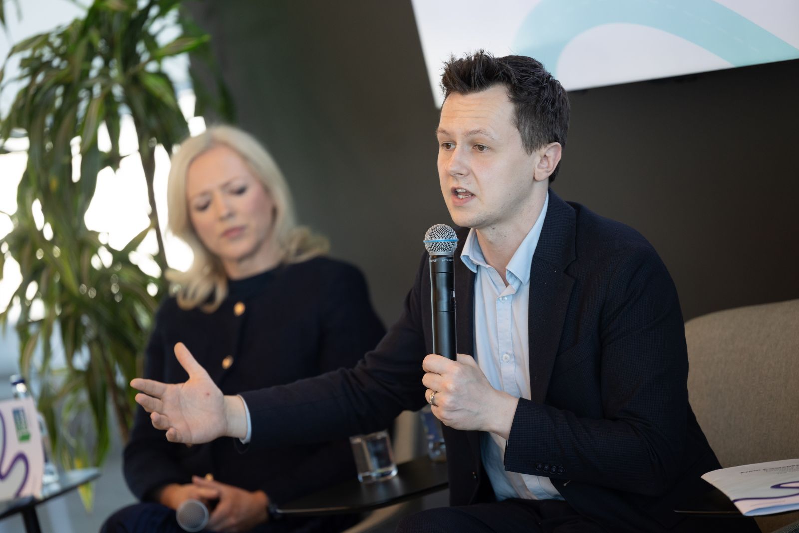 Man wearing dark suit speaking into a microphone, seated blonde woman in the background