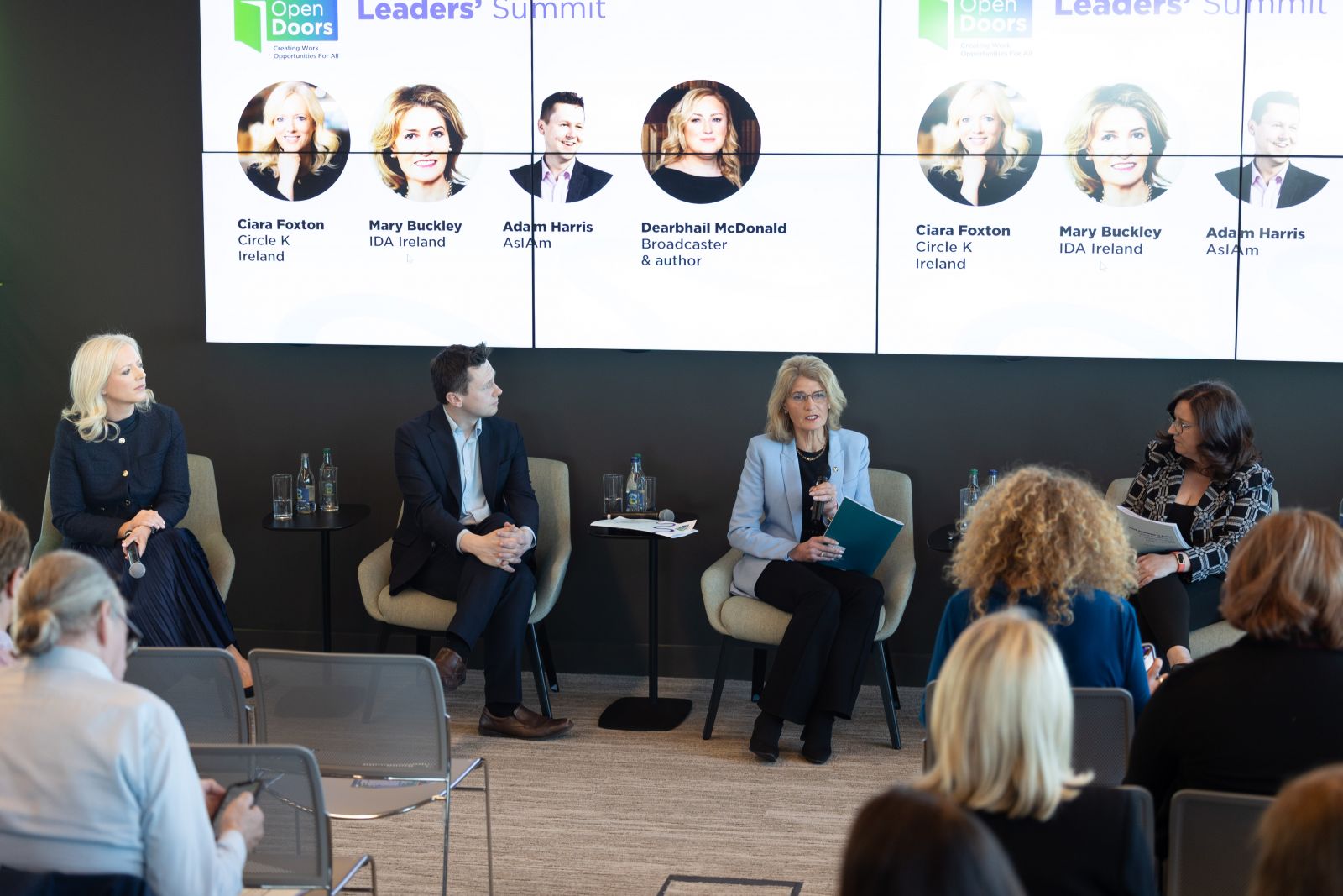 Group of three seated panellists and a moderator, one woman speaking into a microphone, with rear view of seated audience